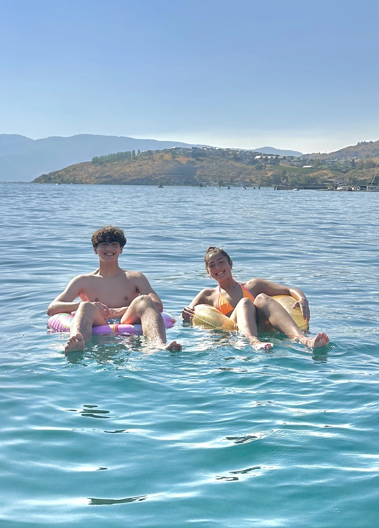 Two friends smiling while floating on inflatable rings in a sunny lake